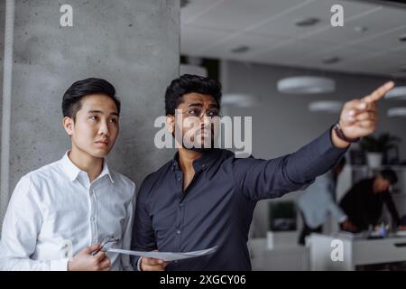 Mix raced couple of male business partners analyzing reports against grey wall. Indian and Chinese men in formal shirts standing in grey office boardr Stock Photo