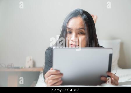 A woman lying on a bed and using a tablet. She appears to be focused on the screen. The background is minimalistic with a small table and some objects Stock Photo