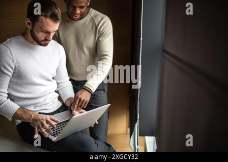 Two concentrated black and white businessmen in casual wear discussing project sitting at office desk, near the window, using pc laptop, looking at sc Stock Photo