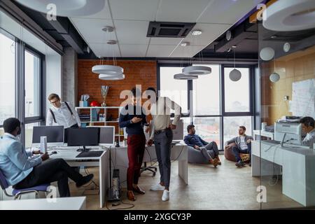 Achieving best results. Two confident young men looking at laptop monitor while their colleagues working in the background Stock Photo