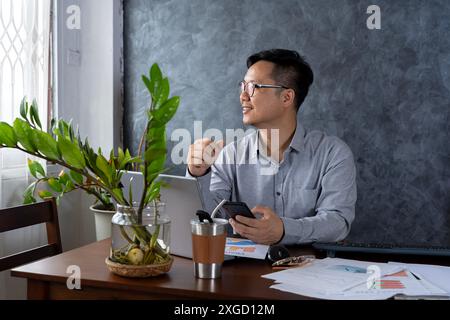 Successful asian man inside office looking out the window, boss in shirt holding smartphone, man in glasses and shirt dreaming about future achievemen Stock Photo