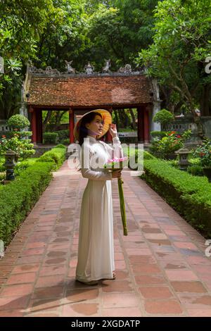 Pretty Vietnamese young woman wearing traditional Ao Dai tunic holding bunch of lotus flowers at Temple of Literature, Hanoi, North Vietnam, Asia Stock Photo