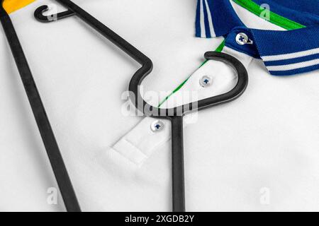 White polo shirt with a blue green collar and yellow sleeves and a black plastic hanger lying on top. Stylish casual sportswear. Copy space Stock Photo