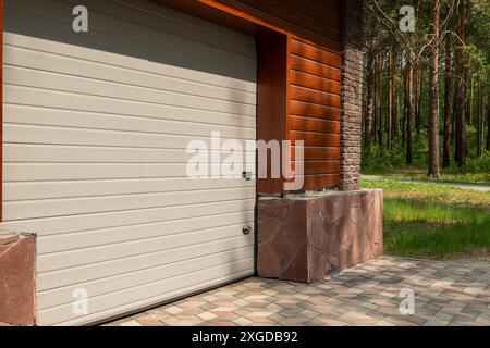 Modern garage door with horizontal panels and keypad beside a dark brown brick wall. Light color contrast against dark brick with lush green trees in Stock Photo