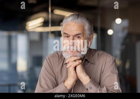 Close-up of a thoughtful senior man with gray beard and headphones, sitting in an office environment. The image captures a moment of reflection and concentration. Stock Photo