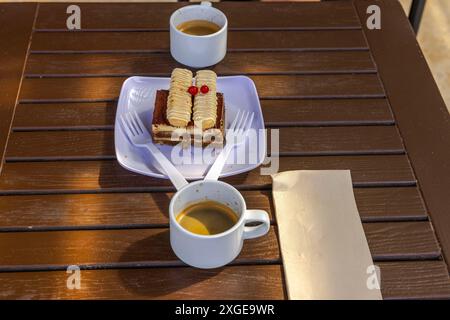 Two cups of coffee and dessert with cream and berries on white plate, placed on wooden table with two plastic forks and napkin. Stock Photo