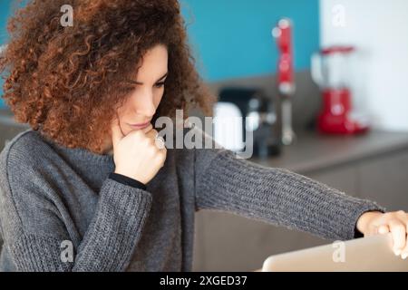Concentrated and thoughtful, a curly-haired woman sits in her kitchen, deeply engaged with her laptop. Her posture and serious expression indicate she Stock Photo