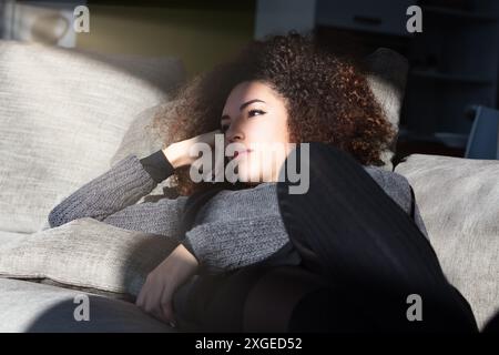 Curly-haired woman lounges on her couch in the living room, lost in thought. Her introspective expression indicates she's reflecting on profound and w Stock Photo