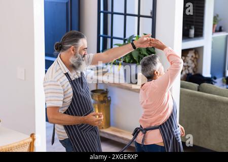 Dancing together, senior couple enjoying time in kitchen wearing aprons Stock Photo