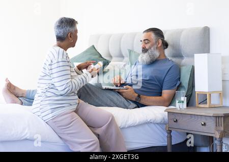 Senior woman giving medication to man using laptop while resting in bed Stock Photo