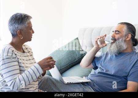 Senior man drinking water while using laptop, talking with woman on bed Stock Photo