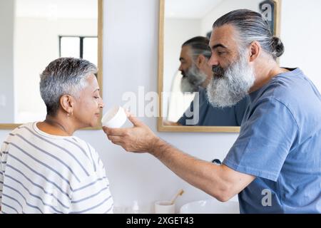 Applying face cream, senior man helping woman in bathroom, morning routine Stock Photo