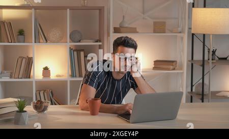 Boring work. Tired freelancer. Frustrated man typing on laptop to meet deadline sitting at desk with cup of coffee in modern home office interior. Stock Photo