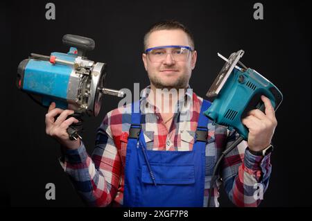 Caucasian repairman in goggles holding woodworking tools. Carpenter portrait on black studio background Stock Photo