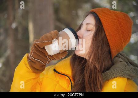 Attractive caucasian young woman in a hat and down jacket drinks hot coffee or tea from a paper cup in a winter snowy forest Stock Photo