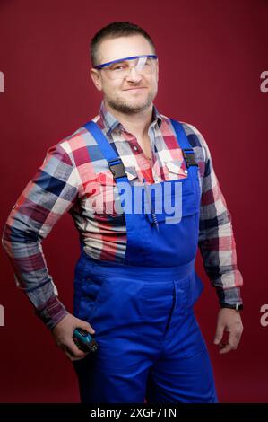 Portrait of a positive friendly Caucasian male foreman repairman in overalls and goggles on a dark red background. Smiling looking into the camera. Stock Photo