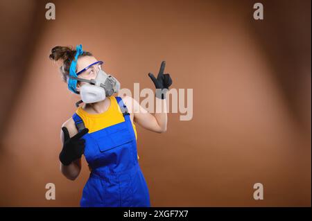 A young athletic caucasian woman, in construction overalls, a respirator, goggles and gloves, stands on a colored background. layout for mock up Stock Photo