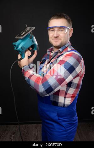 Caucasian repairman in goggles holding woodworking tools. Carpenter portrait on black studio background Stock Photo