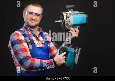 Portrait of smiling young male builder holding tools in both hands while standing and looking at camera on black background Stock Photo