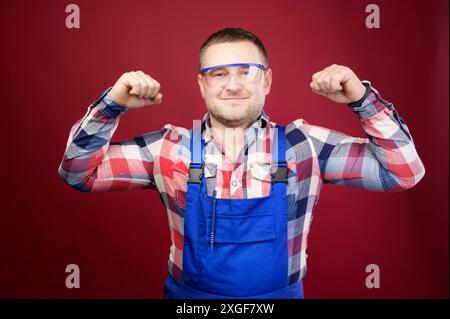 Portrait of a positive friendly Caucasian male foreman repairman in overalls and goggles on a dark red background. Smiling looking into the camera. Stock Photo