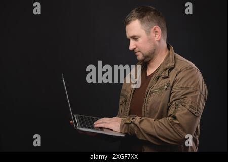 Portrait of an optimistic respectable man in a shirt, standing with a laptop in his hand, looking at the display and working. Studio shot on black Stock Photo