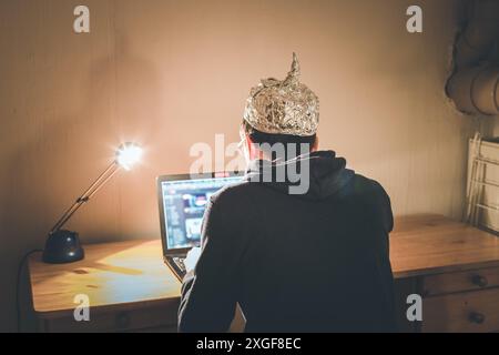 Young man with aluminum cap is sitting in the dark basement in front of a laptop. Conspiracy theory concept Stock Photo