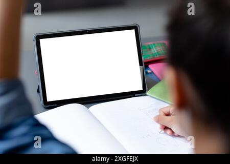 Biracial schoolgirl sitting at desk writing in class and using tablet, with copy space on screen Stock Photo