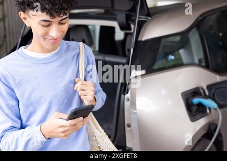 Charging electric car, man using smartphone and carrying reusable shopping bag Stock Photo