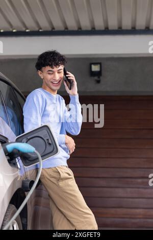 Talking on smartphone, man leaning on electric car while charging at home, copy space Stock Photo