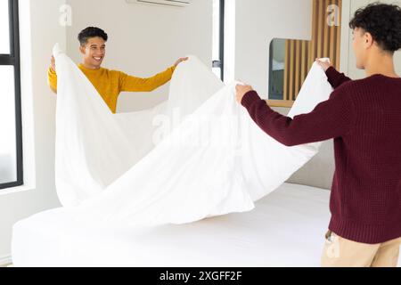 Making bed together, male gay couple smiling and holding white sheet in bedroom Stock Photo