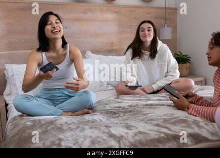 Laughing and chatting, three female friends using smartphones on bed at home Stock Photo