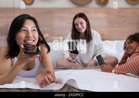 Using smartphones, three women friends relaxing and chatting on bed at home Stock Photo