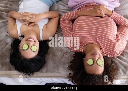 Relaxing at home, two women friends lying on bed with cucumber slices on eyes Stock Photo