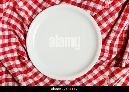 Empty white plate on checkered plaid red and white tablecloth from above as background, top view Stock Photo