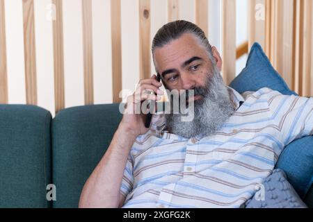 Talking on smartphone, senior man with beard sitting on couch indoors Stock Photo