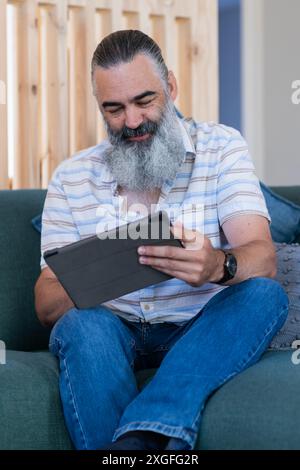 Using tablet, senior man with gray beard sitting on couch, smiling and reading Stock Photo
