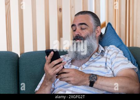 Using smartphone, bearded senior man relaxing on couch, checking messages Stock Photo