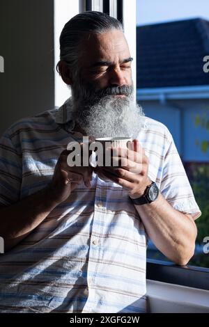 Holding coffee cup, senior man with beard enjoying morning sunlight by window Stock Photo