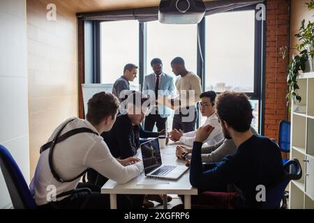 deadline, technology and people concept - creative team of multiracial men, sitting round the desk in diverse casual and formal clothes talking over n Stock Photo