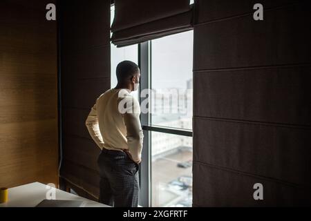 Serious dark skinned businessman, feeling worried about new business project, dressed in knitted white pullover, spends free time at office, looking a Stock Photo