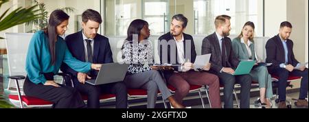 Group of young business people sitting on chairs in row with resumes and laptops and talking. Stock Photo