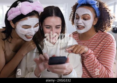 Applying face masks and using smartphone, three women friends enjoying skincare routine Stock Photo