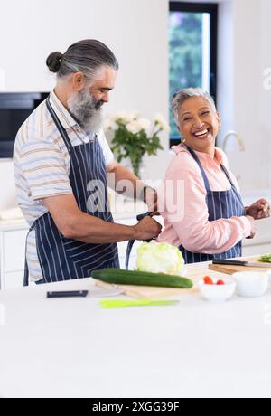 Senior couple in kitchen, man tying apron on woman, preparing to cook together Stock Photo