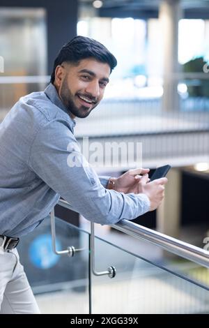 Smiling businessman leaning on railing and holding smartphone in modern office Stock Photo