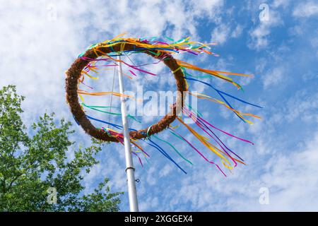 A maypole with colorful ribbons adorning it stands tall against a backdrop of blue sky and white clouds. Stock Photo