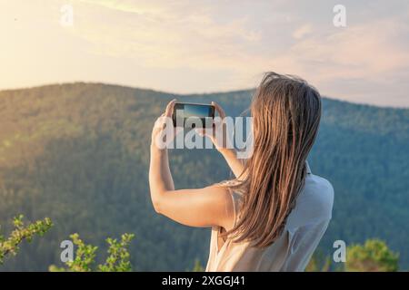 A woman is capturing a beautiful mountain landscape using her smartphone in a peaceful outdoor setting Stock Photo