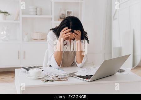 Desperate young woman holds head in hands, feeling overwhelmed by financial paperwork at white office desk, struggling with bills, taxes, and credit Stock Photo