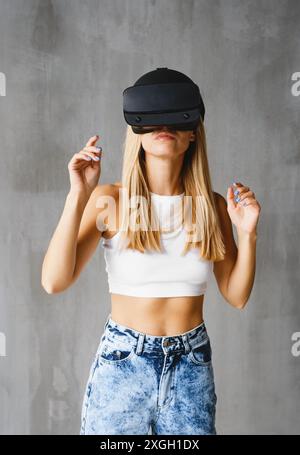 A woman wearing a VR headset interacts with the virtual environment. person explores the digital world. over grey wall Stock Photo