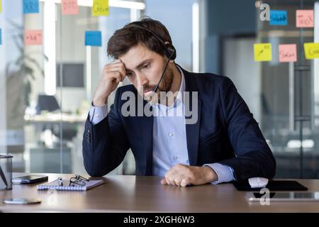 A businessman wearing a suit and headset appears frustrated while working at his desk in a modern office. Sticky notes are visible in the background, indicating a busy and stressful work environment. Stock Photo