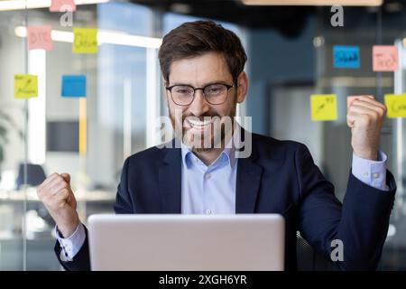 Cheerful businessman in a suit celebrating a triumph while working on a laptop in a modern office. The room features glass walls adorned with colorful sticky notes. Energetic and successful business Stock Photo